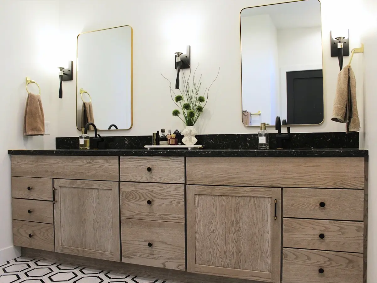 Double sink vanity with light wood finish and black countertop, accented by gold mirrors and lighting.
